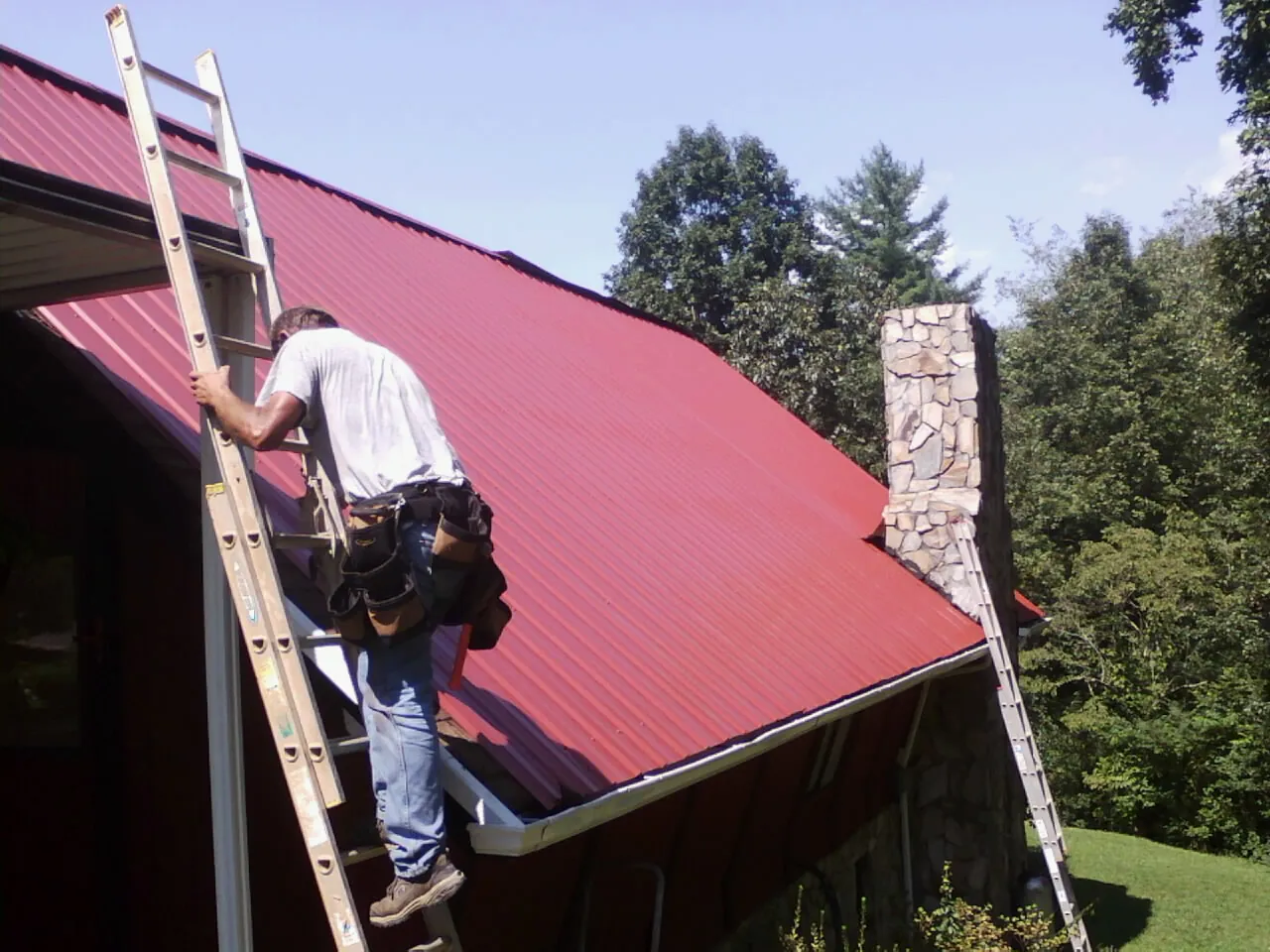Roofer on a ladder beside a red metal roof and stone chimney — Soffit & Fascia Repair by Roofing Brothers in Truxton